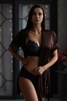 Self assured young alluring lady with long dark hair taking off silk robe and standing in classic styled decorated room in various sexy lingerie during Christmas holidays at home