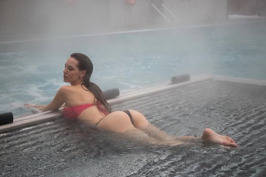 Sexy barefoot female in red swimsuit lying on grates in water of hot spring on resort