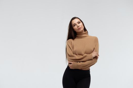 Attractive slim lady in beige jacket and leggings embracing body and looking at camera against white background