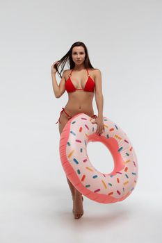 Full body of young slim female model in sexy red bikini carrying donut colored swimming ring and touching hair against white background