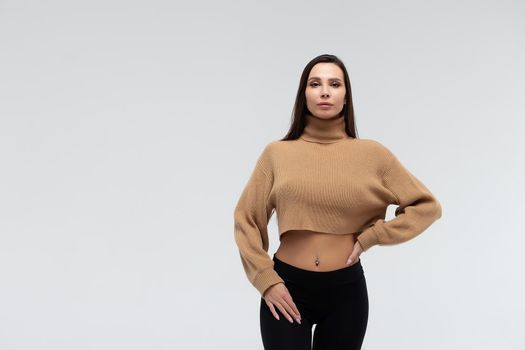 Attractive slim lady in beige jacket and leggings embracing body and looking at camera against white background