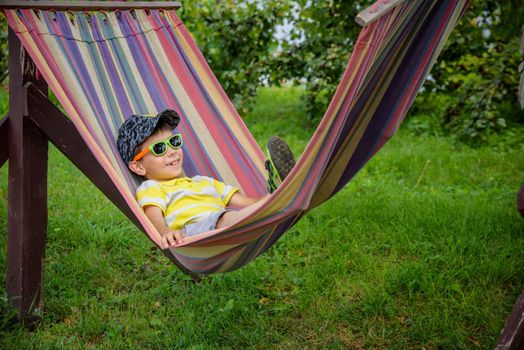 Cute little Caucasian boy relaxing and having fun in multicolored hammock in backyard or outdoor playground. Summer active leisure for kids. Child swinging on hammock. Activities for children.