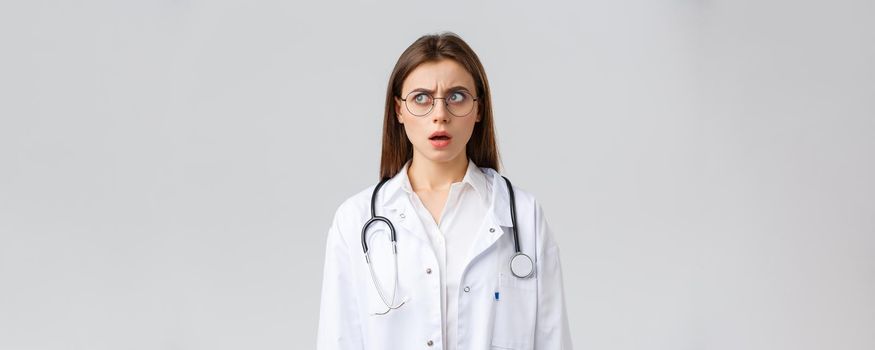 Healthcare workers, medicine, insurance and covid-19 pandemic concept. Shocked and confused young female doctor in white scrubs and glasses, stethoscope, stare left with concerned nervous face.