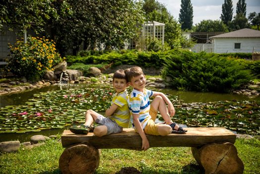 Two brother boys sitting on a bench in a park near peaceful lake with water lily Nymphaeum. Kids relaxing on nature on hot summer day.