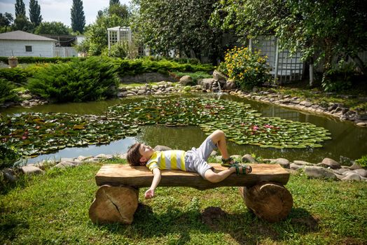 Boy is laying on a bench in a park near peaceful lake. Kid relaxing on nature on hot summer day. Clear blue sky.