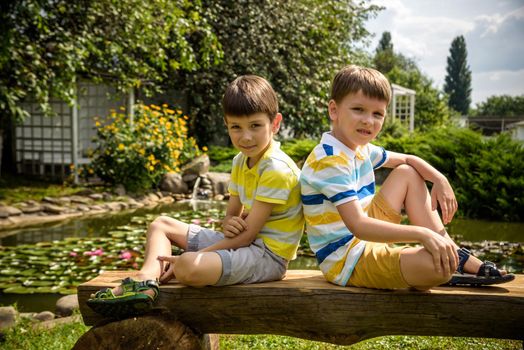 Two brother boys sitting on a bench in a park near peaceful lake with water lily Nymphaeum. Kids relaxing on nature on hot summer day.