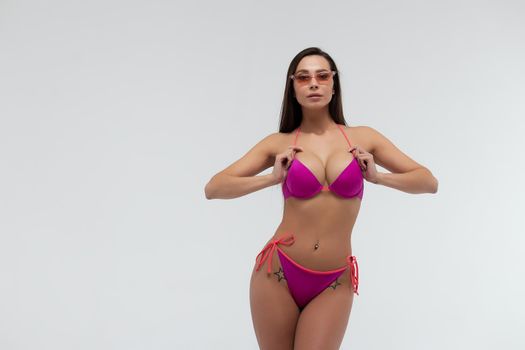 Full body of young slim brunette with perfect sexy body dressed in red bikini and sunglasses standing against white background