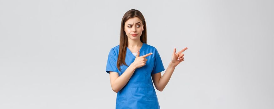 Healthcare workers, prevent virus, insurance and medicine concept. Skeptical and doubtful female nurse or doctor in blue scrubs, smirk and stare judgemental, pointing fingers right.