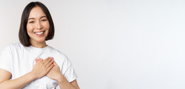 Close up of smiling korean woman holding hands on heart, care and love concept, feel affection, tenderness or heartwarming feeling, standing over white background.