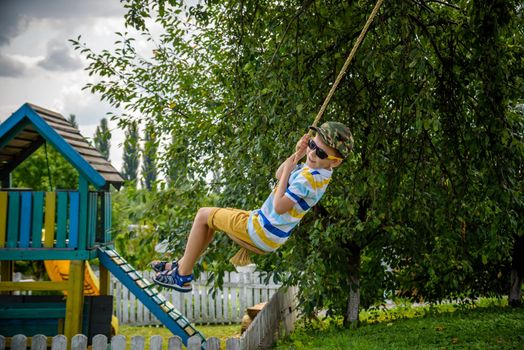 Happy little boy is having fun on a rope swing which he has found while having rest outside city. Active leisure time with children.