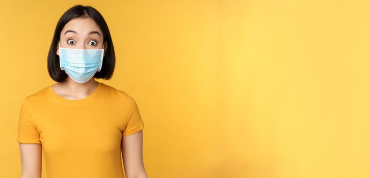 Portrait of korean girl in medical face mask looking surprised, amazed reaction to news, standing over yellow background.