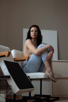 Sensual young female in jeans covering naked breast with hand while relaxing alone in chair in cozy room at home