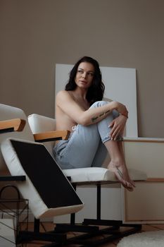 Sensual young female in jeans covering naked breast with hand while relaxing alone in chair in cozy room at home