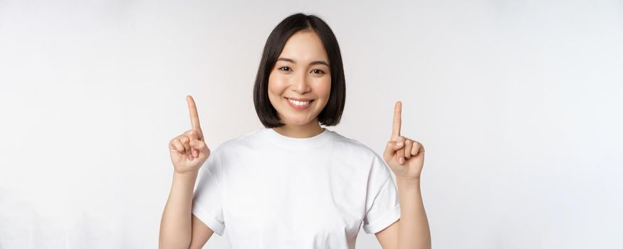 Portrait of enthusiastic young woman, asian girl smiling pointing fingers up, showing advertisement upwards, standing over white background.