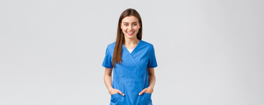 Healthcare workers, prevent virus, insurance and medicine concept. Cheerful smiling beautiful doctor, female nurse in blue scrubs, intern in hospital staying positive, standing grey background.