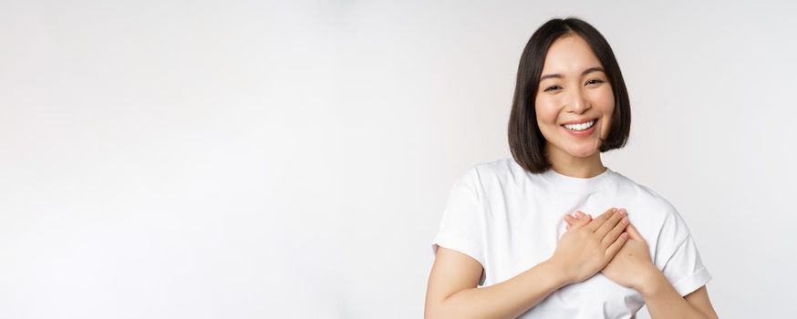 Close up of smiling korean woman holding hands on heart, care and love concept, feel affection, tenderness or heartwarming feeling, standing over white background.