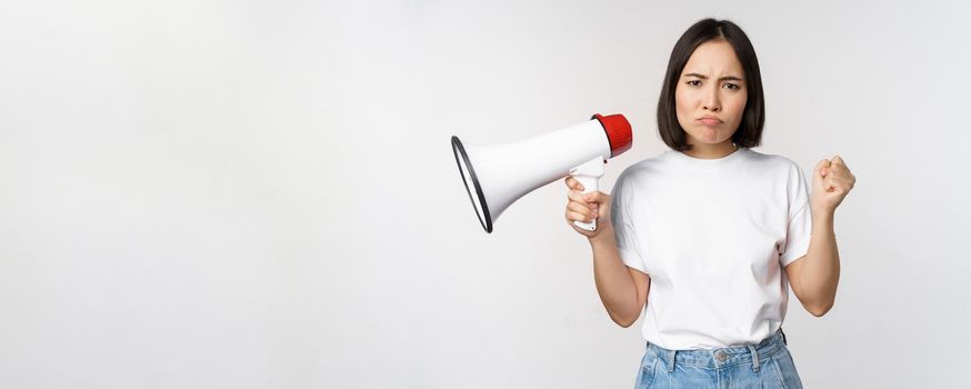 Angry asian girl activist, holding megaphone and looking furious, protesting, standing over white background. Copy space