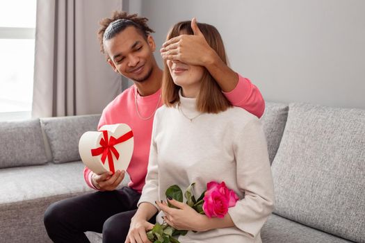 A guy in a pink jumper and a girl in a white dress, on a gray couch . The guy covered the girls eyes with his hand to give a surprise - a white heart-shaped box with a bow and a bouquet of roses. copy space