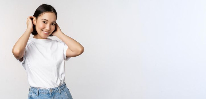 Portrait of cute, beautiful asian woman touching her new short haircut, showing hairstyle, smiling happy at camera, standing over white background.