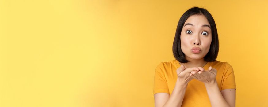 Cute asian girl sending air kiss, blowing mwah with puckered lips, standing over yellow background. Copy space