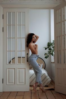 Full body of barefoot young slim female in trendy ripped jeans covering naked breast with hand while leaving bedroom at home