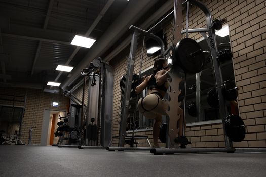 Low angle back view of slim athletic woman in sportswear with perfect sexy buttocks doing squats with heavy barbell during workout in gym