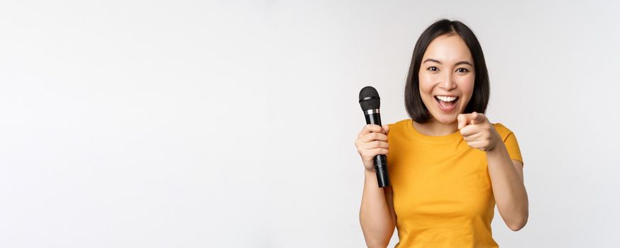 Enthusiastic asian girl with microphone, pointing finger at camera, suggesting you to sing, standing over white background.