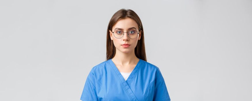 Healthcare workers, medicine, insurance and covid-19 pandemic concept. Young female intern, nurse or doctor in scrubs and glasses, looking at camera with serious expression, grey background.