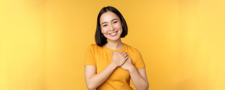 Romantic korean girl, asian woman holding hands on heart, smiling with care and tenderness, standing over yellow background.