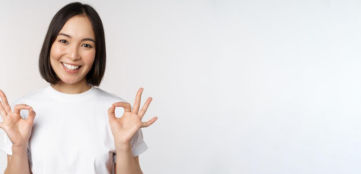 Very well, excellent. Smiling asian woman showing okay sign, approval, ok gesture, looking satisfied, recommending smth, standing over white background.
