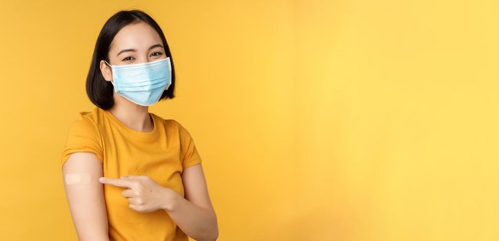 Vaccination and covid-19 pandemic concept. Smiling asian woman in medical face mask, showing her shoulder with band aid after vaccinating from coronavirus, yellow background.