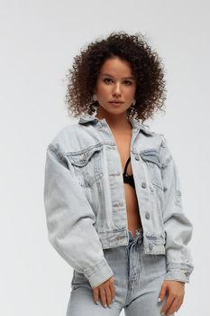 Full body of confident young barefoot female model with curly hairstyle wearing trendy denim jacket and jeans