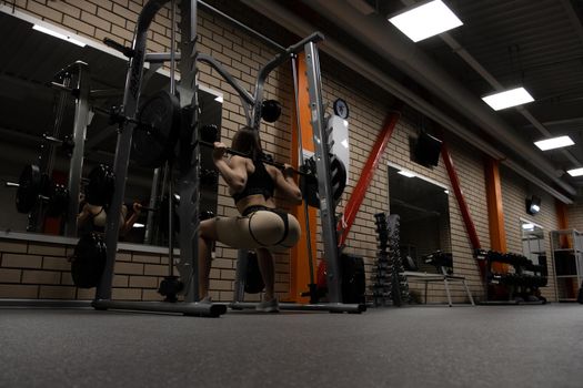 Low angle back view of slim athletic woman in sportswear with perfect sexy buttocks doing squats with heavy barbell during workout in gym