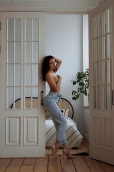 Full body of barefoot young slim female in trendy ripped jeans covering naked breast with hand while leaving bedroom at home