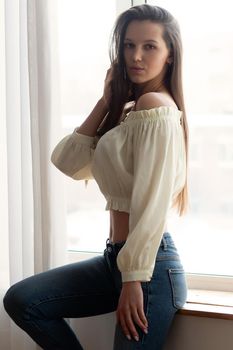 Young skinny female in stylish top and jeans raising arm and leaning on door in light bedroom at home