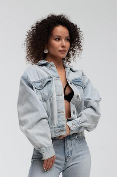 Full body of confident young barefoot female model with curly hairstyle wearing trendy denim jacket and jeans