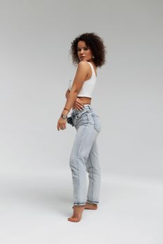 Full body of confident young barefoot female model with curly hairstyle wearing trendy denim jacket and jeans