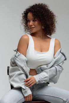 Full body of confident young barefoot female model with curly hairstyle wearing trendy denim jacket and jeans sitting on chair in white studio