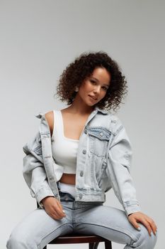 Full body of confident young barefoot female model with curly hairstyle wearing trendy denim jacket and jeans sitting on chair in white studio
