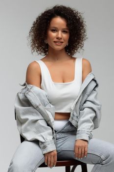 Full body of confident young barefoot female model with curly hairstyle wearing trendy denim jacket and jeans sitting on chair in white studio