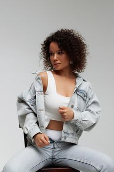 Full body of confident young barefoot female model with curly hairstyle wearing trendy denim jacket and jeans sitting on chair in white studio