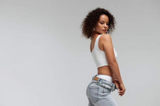 Full body of confident young barefoot female model with curly hairstyle wearing trendy denim jacket and jeans