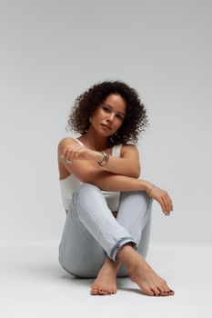 Full body of confident young barefoot female model with curly hairstyle wearing trendy denim jacket and jeans