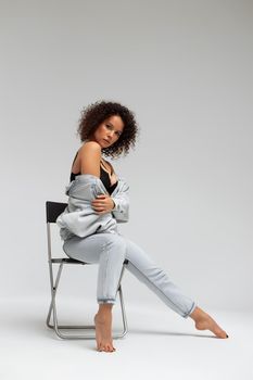 Full body of confident young barefoot female model with curly hairstyle wearing trendy denim jacket and jeans sitting on chair in white studio