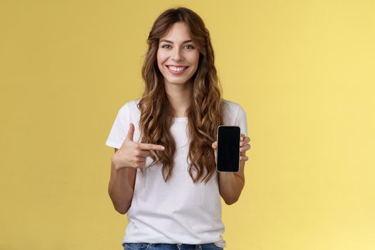 Girl have everything need smartphone. Cheerful relaxed carefree woman long curly hairstyle hold mobile phone pointing forefinger cellphone smiling broadly explain how app works yellow background.