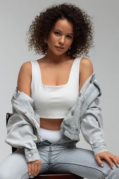 Full body of confident young barefoot female model with curly hairstyle wearing trendy denim jacket and jeans sitting on chair in white studio