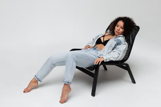 Full body of confident young barefoot female model with curly hairstyle wearing trendy denim jacket and jeans sitting on chair in white studio