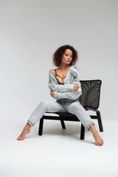 Full body of confident young barefoot female model with curly hairstyle wearing trendy denim jacket and jeans sitting on chair in white studio