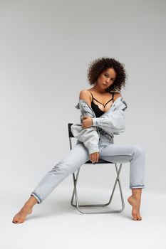 Full body of confident young barefoot female model with curly hairstyle wearing trendy denim jacket and jeans sitting on chair in white studio