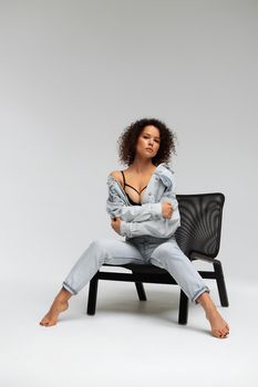 Full body of confident young barefoot female model with curly hairstyle wearing trendy denim jacket and jeans sitting on chair in white studio